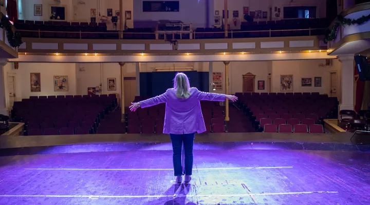 Devyn stands on center stage at the Abbeville Opera House. Her arms stretched out to the side and facing an empty theater. She is illuminated by purple stage lighting.