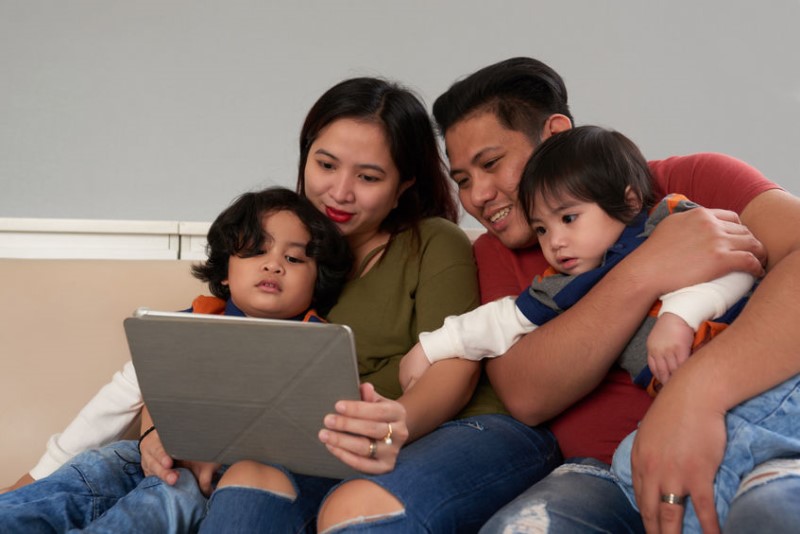 photo of mom & dad sitting on couch and holding their two young children while they all look at a tablet screen together