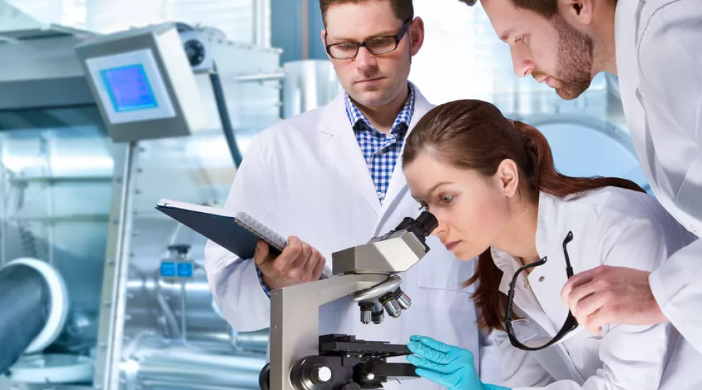 3 scientists in a lab, one looking through a microscope