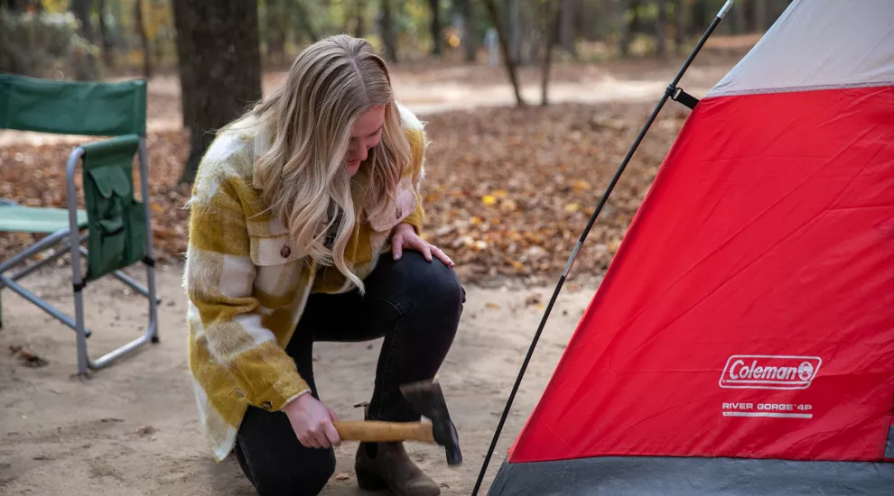 Devyn, wearing a yellow and white plaid shirt jacket with dark blue jeans kneels beside a red and gray camping tent while holding a hatchet.