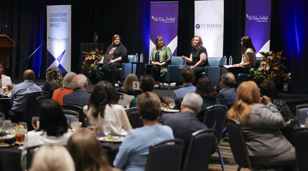 photo of WhatWorksSC Award Celebration showing a seated crowd, seated panelists on a stage and Riley Institute and Furman banners in the background