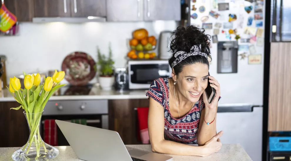 picture of woman on phone in her kitchen with flowers and a laptop in the background