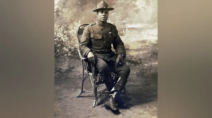 Unidentified African American soldier in uniform with marksmanship qualification badge and campaign hat, with cigarette holder in front of painted backdrop.