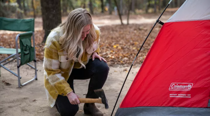 Devyn, wearing a yellow and white plaid shirt jacket with dark blue jeans kneels beside a red and gray camping tent while holding a hatchet.