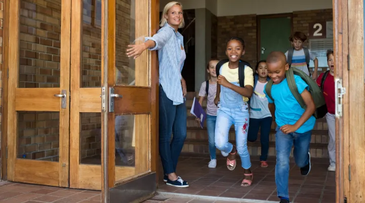 photo of teacher holding school door open as students run out excitedly