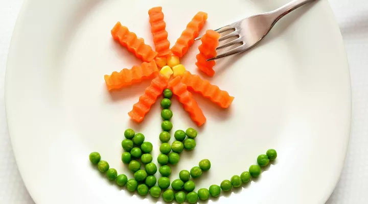 Peas, carrots, and corn arranged on a plate to look like a flower. A fork reaches for one of the carrots. 